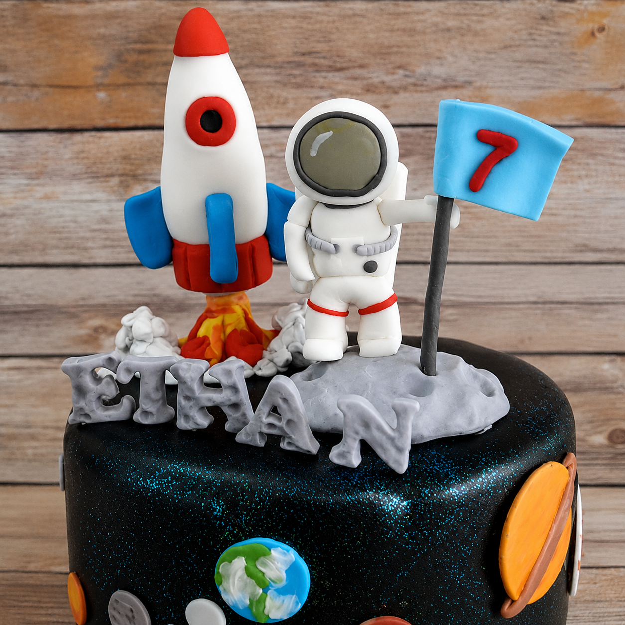 Edible space-themed cake decorating kit with a white-and-red rocket, a white astronaut holding a blue number 7 flag, gray Ethan letters, and colorful planets.