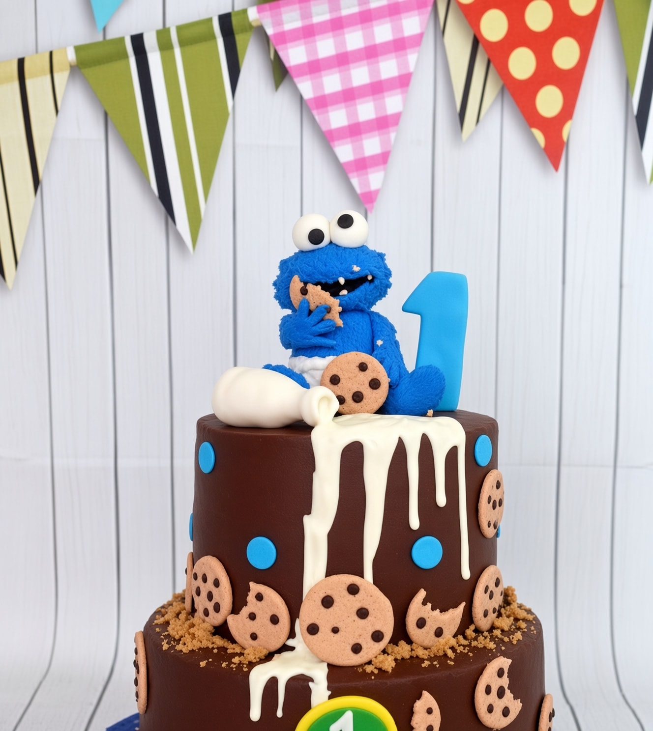 Two-tier chocolate cake decorated with blue dots, cookie details, white icing drips, and an edible blue Cookie Monster-style topper with milk and cookies.